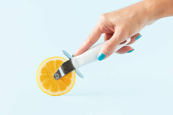 Female hand holds pizza cutter with lemon slice on isolated pastel blue background. Healthy lifestyle and supplements as a sources of vitamin C. Minimal aesthetic abstract concept of boost immunity. Health care and Illness prevention. - Foto, Imagem