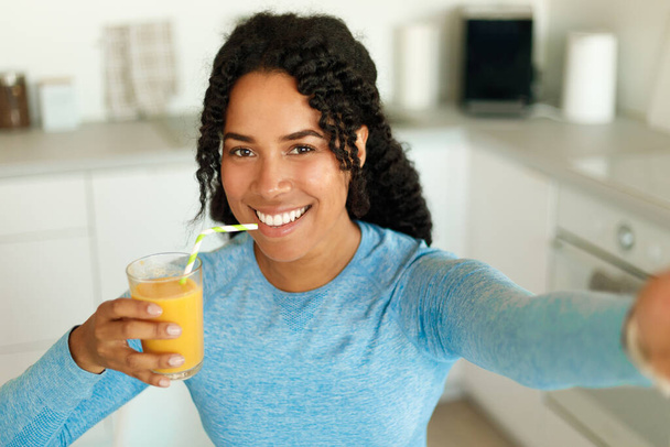Positive african american fit woman drinking fresh juice from straw and taking selfie, standing in kitchen interior. Happy black lady in sportswear resting after domestic workout - Foto, Imagen