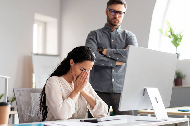 Strenge Millennial European Guy Chef Blick auf weinende Managerin am Arbeitsplatz mit Computer, finden Fehler in der modernen Büroeinrichtung. Stress, Druck, Probleme in Unternehmen, Finanzen, Arbeit im Unternehmen - Foto, Bild