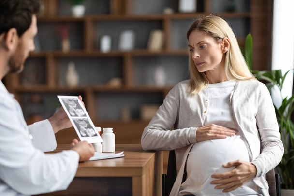 Mies Gynekologi lääkäri näyttää vauvan sonografia kuva huolissaan raskaana oleva nainen kokouksen aikana toimistossa, ammatillinen lisääntymiseen endokrinologi selittää seulonta tulos naispotilaalle - Valokuva, kuva