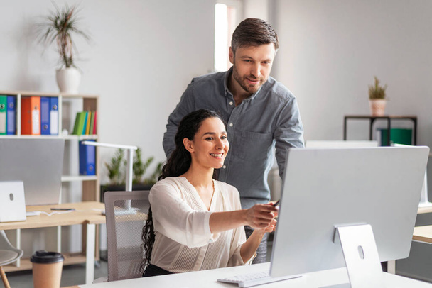 Smiling millennial european female showing to man manager with hand in computer screen in modern office interior. Business and teamwork, finance, data analysis, developing in company and planning - 写真・画像