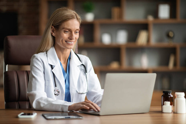 Muotokuva hymyilevä nainen lääkäri univormussa työskentelee kannettavan tietokoneen toimistossa, kaunis lääkäri nainen yllään Medical Coat Consulting potilaiden verkossa tai selaamalla Internetiä, Kopioi tilaa - Valokuva, kuva