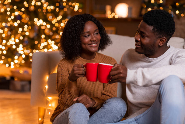 Boldog fiatal afro-amerikai pár éljenzés csésze forró ital, élvezze a nyaralást és a szabadidőt, ülni a padlón a nappali belső világító fények. Ünnepnap, este kakaóval otthon - Fotó, kép