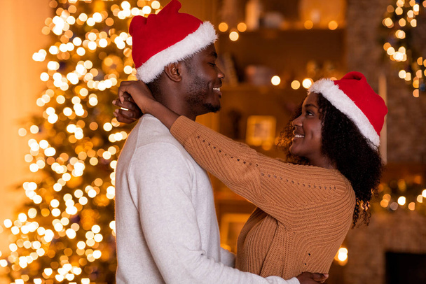 Hymyilevä tuhatvuotinen musta mies ja nainen Santa hatut halailemassa pimeässä huoneessa joulukuusi valoisa seppeleitä. Juhli lomaa ja uutta vuotta yhdessä kotona, rakkautta ja romantiikkaa covid-19: n vuoksi - Valokuva, kuva