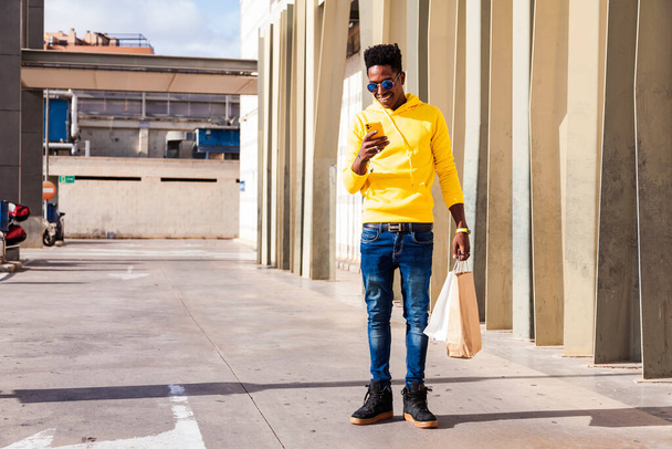 Un joven negro vestido con una sudadera amarilla, vaqueros azules y gafas de sol usando su teléfono inteligente mientras sostiene bolsas de compras. Está fuera de un centro comercial.. - Foto, Imagen