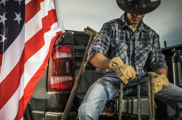 Kaukasischer Cowboy-Rancher in seinen Vierzigern, der auf seinem Pickup-Ladebett sitzt und ein Seil in seinen Händen hält. Werkzeuge für einen Ranch-Job vorbereiten. Amerikanische Flagge neben ihm. - Foto, Bild