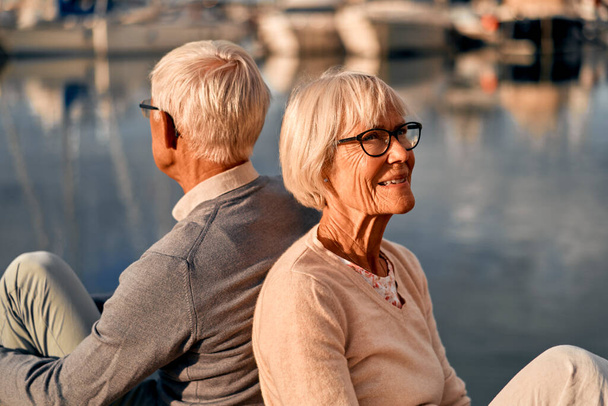 Una pareja de ancianos, inteligentes y canosos enamorados están sentados en un muelle en un puerto marítimo, pasando un momento romántico admirando el paisaje marino. - Foto, imagen