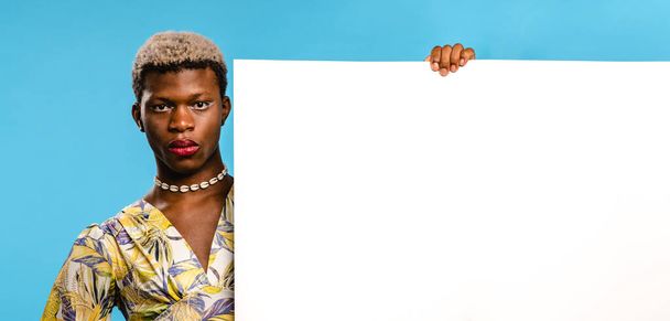 Stylish African transgender woman with dyed hair and makeup in colorful shirt and necklace standing in studio near blue background and holding white blank sheet - Photo, Image