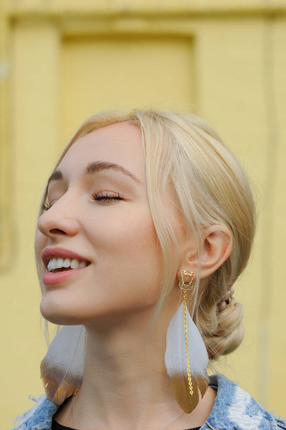 Close-up portrait of gorgeous blonde woman in boho chic feather earrings and jean jacket, bohemian jewelry - Photo, image