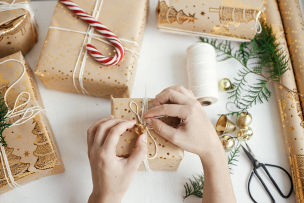 Hands wrapping stylish christmas gifts flat lay. Person preparing modern christmas gift boxes, festive paper, golden ornaments and thread composition on white wood. Atmospheric time, Happy Holidays! - Foto, Bild