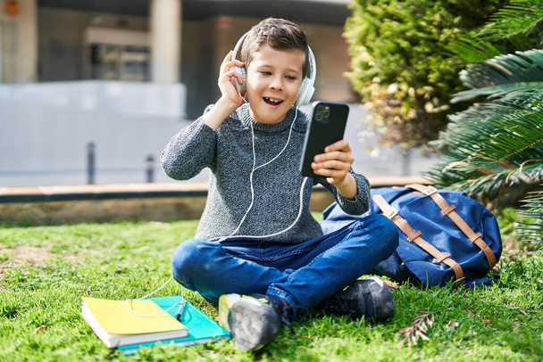Rubio niño estudiante sonriendo confiado escuchando música en el parque - Foto, imagen
