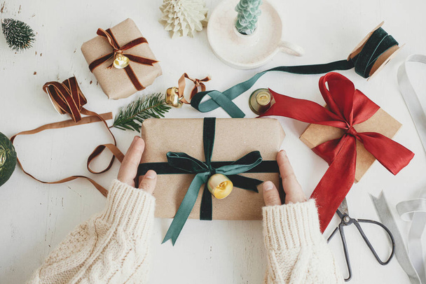 Veselé Vánoce! Ruce balící stylové vánoční dárek byt ležel. Osoba připravující vánoční dárek se zelenou stuhou a ozdobnou kompozicí na bílém dřevě. Estetické zimní prázdniny - Fotografie, Obrázek