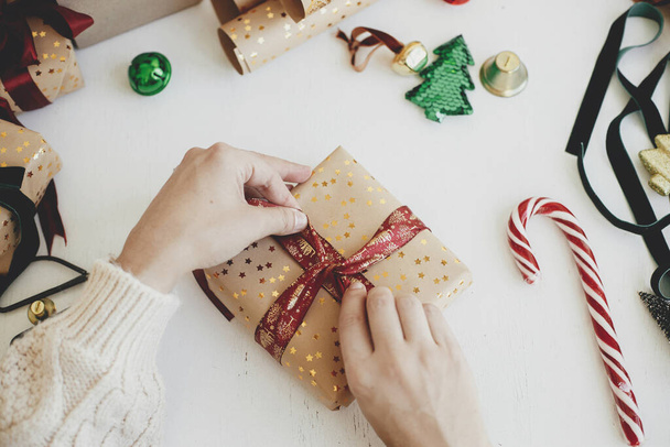 Joyeux Noël ! Mains enveloppant élégant cadeau de Noël plat laïc. Personne préparant boîte cadeau moderne avec ruban rouge, papier d'emballage doré, ornements sur fond blanc. Heure d'hiver dans l'atmosphère - Photo, image