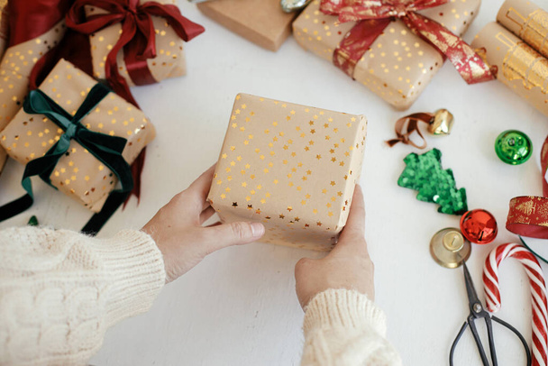 Kezek csomagolása stílusos karácsonyi ajándék. Személy elkészítése modern ajándék doboz arany csomagolópapír, szalagok, díszek fehér háttér. Boldog karácsonyt! Légköri téli időszámítás - Fotó, kép