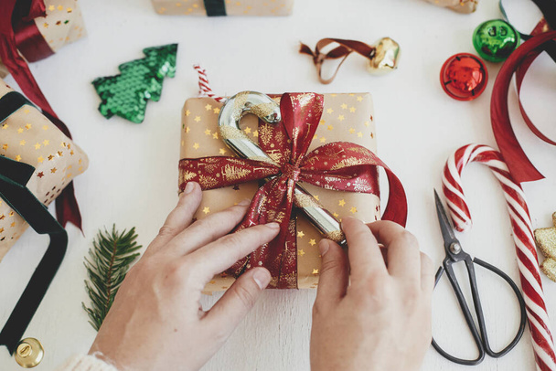 Vrolijk kerstfeest! Handen versieren stijlvolle kerstcadeau. Persoon bereiden moderne geschenkdoos met suikerriet, rood lint, inpakpapier, ornamenten op witte achtergrond. Sfeervolle wintertijd - Foto, afbeelding