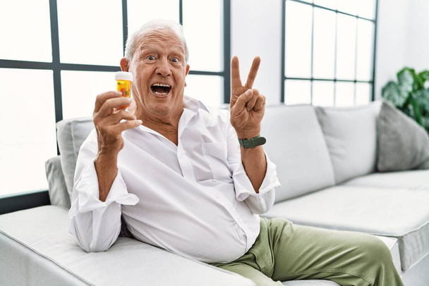 Senior man holding pills smiling with happy face winking at the camera doing victory sign with fingers. number two.  - Photo, Image