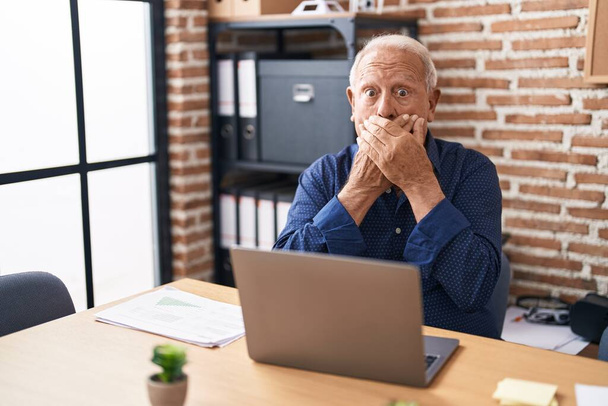 Starszy mężczyzna z siwymi włosami, pracujący przy laptopie komputerowym w biurze, zszokowany zakryciem ust rękami przez pomyłkę. tajna koncepcja.  - Zdjęcie, obraz