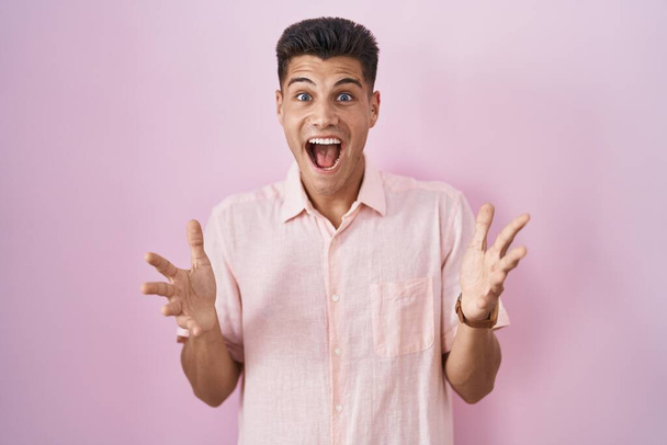 Joven hombre hispano de pie sobre fondo rosa celebrando loco y sorprendido por el éxito con los brazos levantados y los ojos abiertos gritando emocionado. concepto ganador  - Foto, imagen