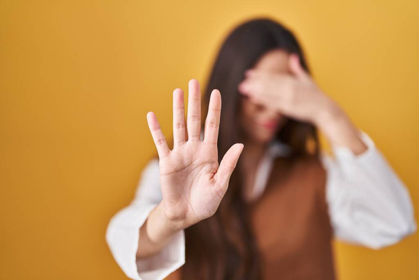 Young brunette woman standing over yellow background wearing glasses covering eyes with hands and doing stop gesture with sad and fear expression. embarrassed and negative concept.  - Fotografie, Obrázek