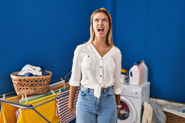 Young blonde woman at laundry room angry and mad screaming frustrated and furious, shouting with anger. rage and aggressive concept.  - Photo, Image