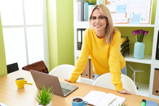 Giovane donna bionda business worker sorridente fiducioso in piedi sulla scrivania in ufficio - Foto, immagini