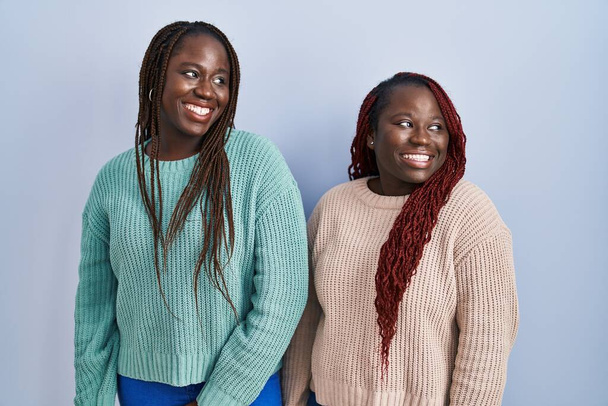 Two african woman standing over blue background looking away to side with smile on face, natural expression. laughing confident.  - Фото, зображення