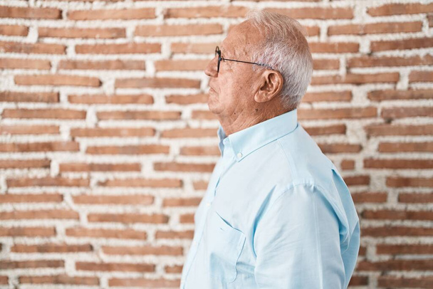 Senior man with grey hair standing over bricks wall looking to side, relax profile pose with natural face and confident smile.  - Photo, Image