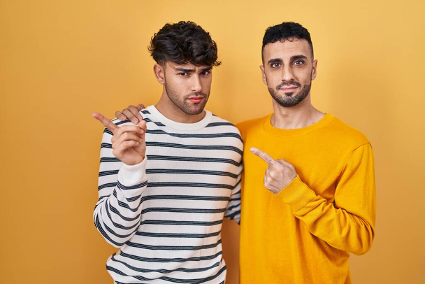 Jeune couple gay hispanique debout sur fond jaune pointant avec le doigt de la main sur le côté montrant la publicité, visage sérieux et calme  - Photo, image