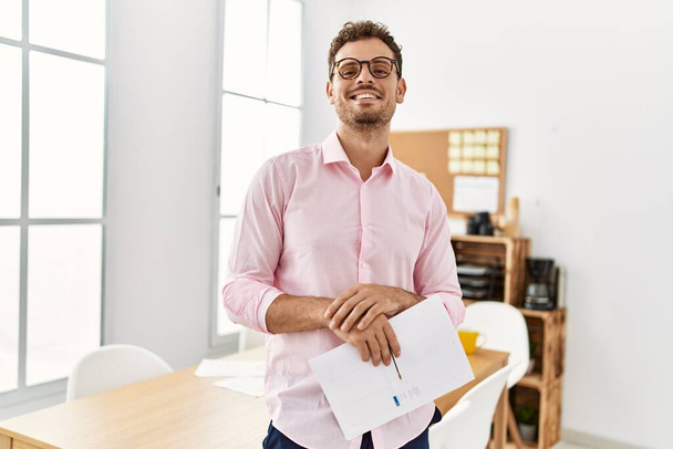 Молодий іспаномовний чоловік посміхається впевнено тримає документ в офісі
 - Фото, зображення