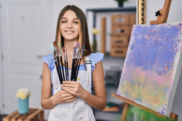 Entzückendes Mädchen lächelt selbstbewusst und hält Pinsel im Kunstatelier - Foto, Bild