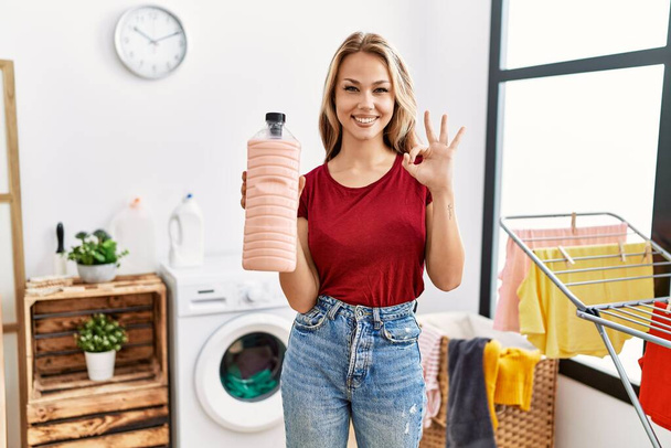 Jonge blanke vrouw met wasmiddel fles in de wasruimte doet ok teken met de vingers, glimlachend vriendelijk gebaar uitstekend symbool  - Foto, afbeelding