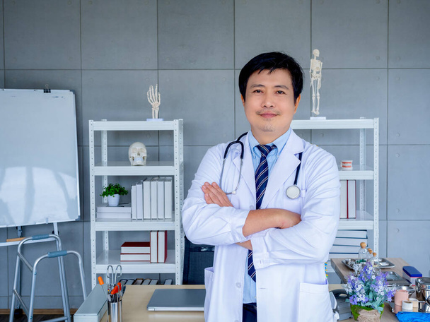 Hymyilevä aasialainen mies ortopedinen lääkäri muotokuva valkoisessa takissa seisoo ristissä kädet lähellä kirjahyllyä ja laitteita lääketieteellisessä toimistossa. Itsevarma aikuinen mies lääkäri tai lääkäri stetoskoopilla. - Valokuva, kuva