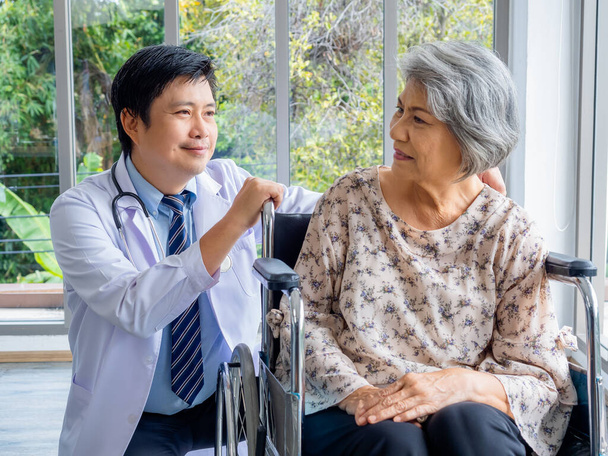 Χαμογελαστός θετικός Ασιάτης γιατρός με λευκό παλτό μιλά με φροντίδα σε ηλικιωμένη ηλικιωμένη ηλικιωμένη ασθενή που κάθεται σε αναπηρικό καροτσάκι στο ιατρικό γραφείο του νοσοκομείου. Φροντίδα, υγειονομική περίθαλψη και ιατρική έννοια. - Φωτογραφία, εικόνα