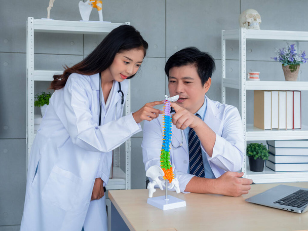 Twee Aziatische artsen in witte uniformen praten en werken samen in het medisch kantoor. De orthopedische volwassen mannelijke arts biedt kennis en begeleiding aan vrouwelijke arts over wervelkolom op bureau in het ziekenhuis. - Foto, afbeelding