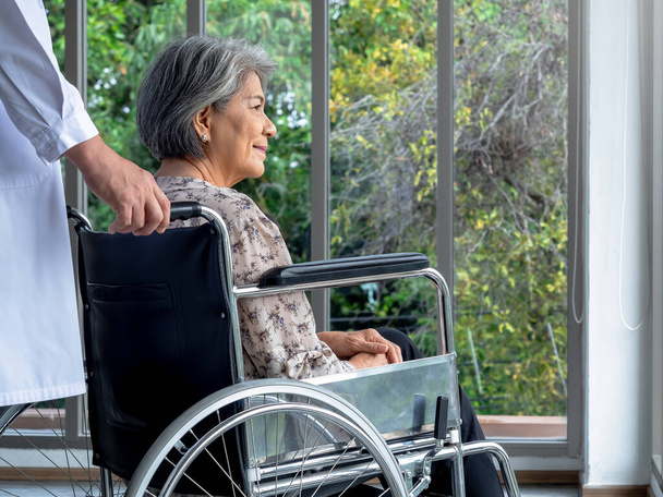 Close-up portret van gelukkige Aziatische bejaarde oudere vrouwelijke patiënt zit in een rolstoel, terwijl de man arts in witte jas verzorgen in het medisch kantoor in het ziekenhuis. Verzorging, gezondheidszorg en medisch concept. - Foto, afbeelding