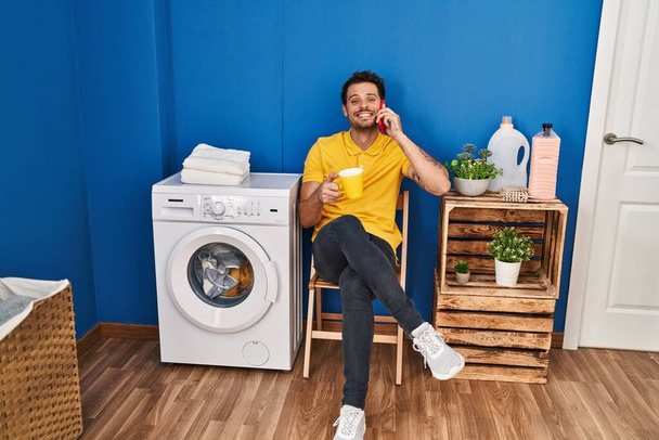 Jovem hispânico bebendo café falando no smartphone wating para máquina de lavar roupa na lavanderia - Foto, Imagem