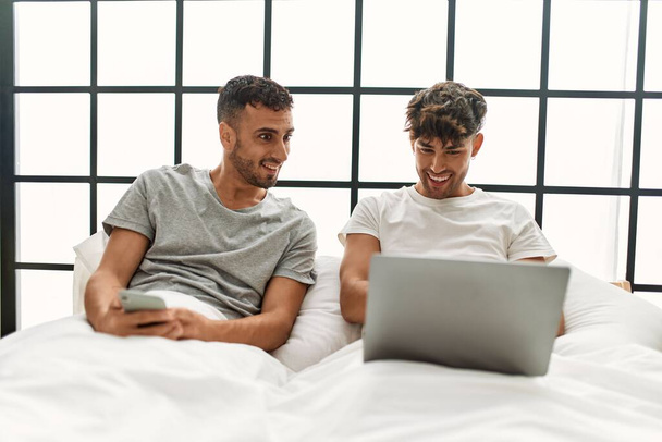 Dwóch Hiszpanów para za pomocą laptopa i smartfona siedzi na łóżku w sypialni - Zdjęcie, obraz