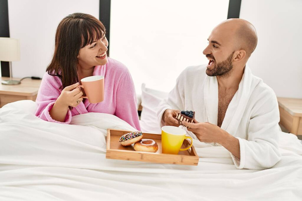 Νεαρό ζευγάρι ισπανόφωνων τρώει πρωινό καθισμένο στο κρεβάτι στο υπνοδωμάτιο. - Φωτογραφία, εικόνα