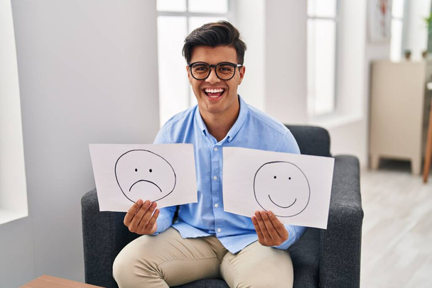 Hombre hispano trabajando en la depresión sosteniendo triste a la emoción feliz papel sonriendo y riendo en voz alta porque broma loca divertida.  - Foto, Imagen