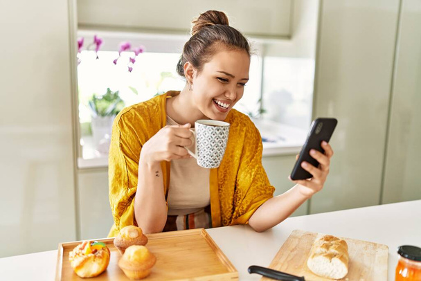 Молодая красивая латиноамериканка завтракает кофе со смартфона на кухне - Фото, изображение