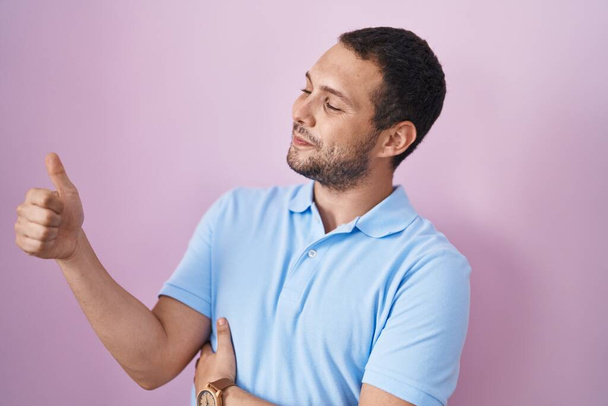 Ισπανόφωνος άντρας στέκεται πάνω από ροζ φόντο, δείχνοντας περήφανος, χαμογελώντας κάνει αντίχειρες επάνω χειρονομία στο πλάι  - Φωτογραφία, εικόνα