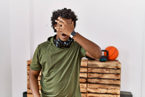 Африканский мужчина с вьющимися волосами в спортивной одежде в спортзале подглядывает в шокирующие закрывающие лицо и глаза рукой, смотрит сквозь пальцы со смущенным выражением лица.  - Фото, изображение