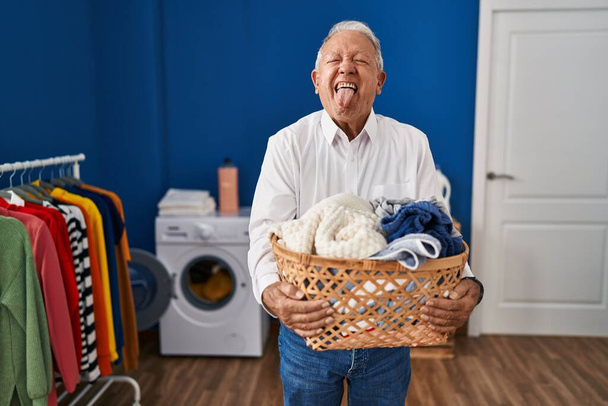Älterer Mann mit grauen Haaren hält Wäschekorb zu Hause und streckt mit lustiger Miene die Zunge heraus.  - Foto, Bild