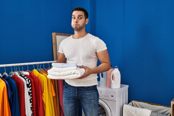 Junger hispanischer Mann mit Bart, der gefaltete saubere Handtücher in der Waschküche hält und mit lustigem Gesicht die Wangen aufbläst. Mund mit Luft aufgeblasen, Luft einfangen.  - Foto, Bild