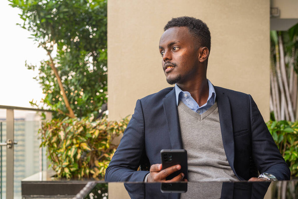 Portrait of handsome black African businessman wearing suit while thinking and using phone - Fotoğraf, Görsel