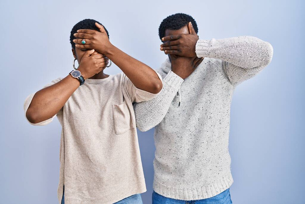 Mladý africký americký pár stojící nad modrým pozadím spolu zakrývající oči a ústa rukama, překvapený a šokovaný. skrývání emocí  - Fotografie, Obrázek