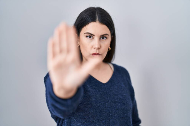 Jonge brunette vrouw die over geïsoleerde achtergrond staan doen stoppen met zingen met palm van de hand. waarschuwingsuitdrukking met negatief en ernstig gebaar op het gezicht.  - Foto, afbeelding