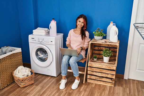 Çamaşır odasında çamaşır makinesi bekleyen beyaz bir kadın dizüstü bilgisayar kullanıyor. - Fotoğraf, Görsel