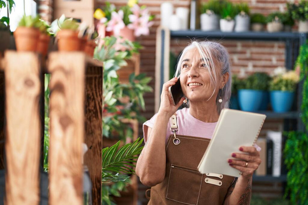 Middle age grey-haired woman florist talking on smartphone reading book at florist - 写真・画像