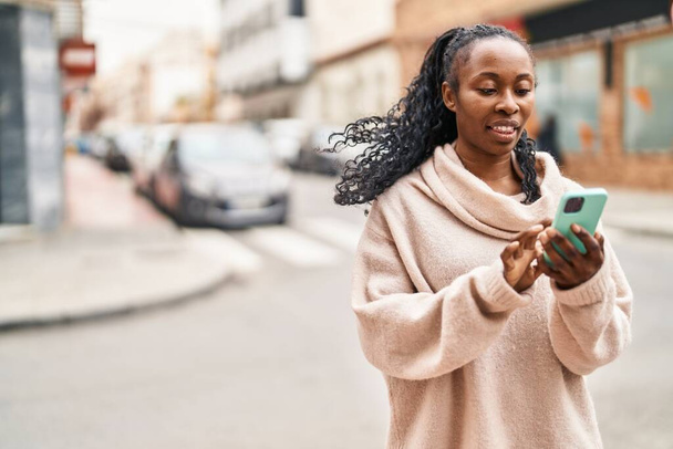 Afrikaans amerikaanse vrouw glimlachen zelfverzekerd met behulp van smartphone op straat - Foto, afbeelding